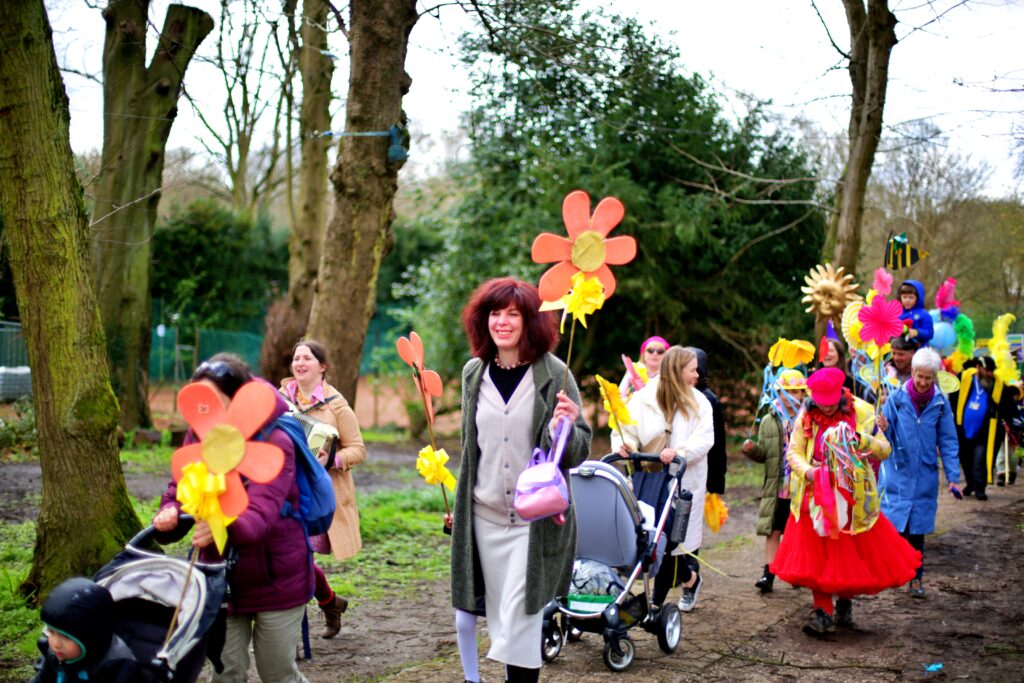 A parade of people of all ages carrying bright flowers, sunshines and bees on sticks moves through Moseley Park, with tress and bushes in the background