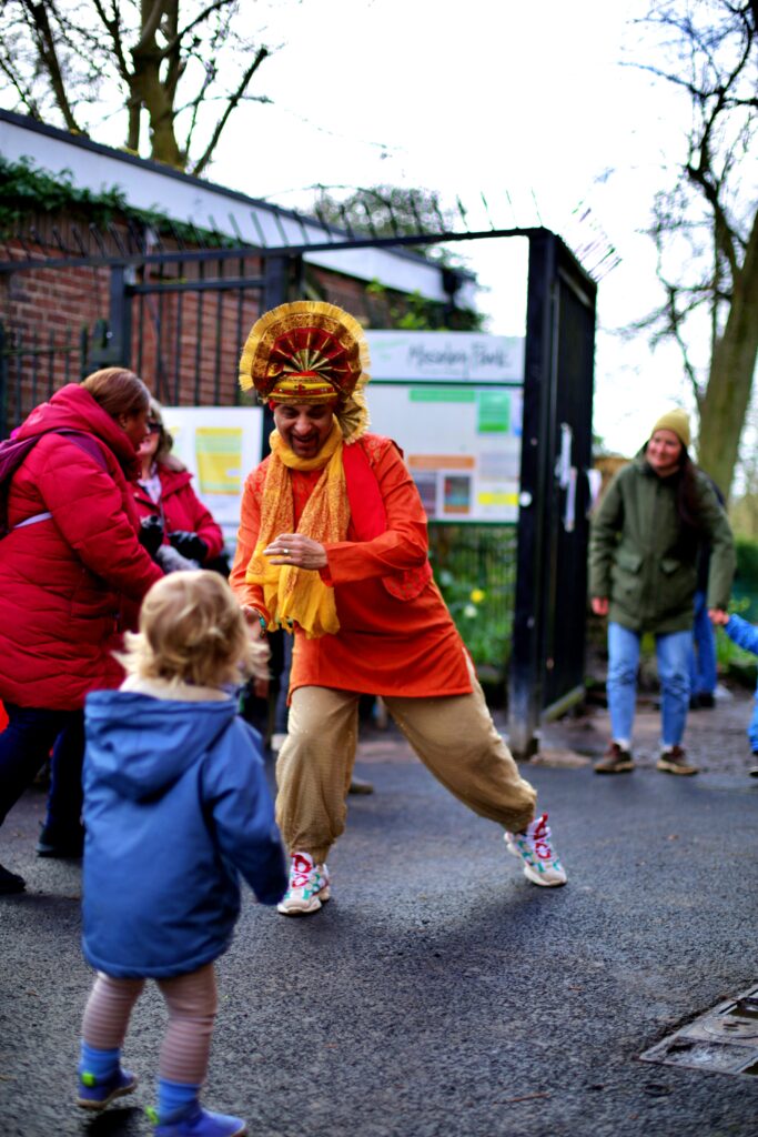 A bhangra dancer in an orange outfit dances at the park gate for a toddler