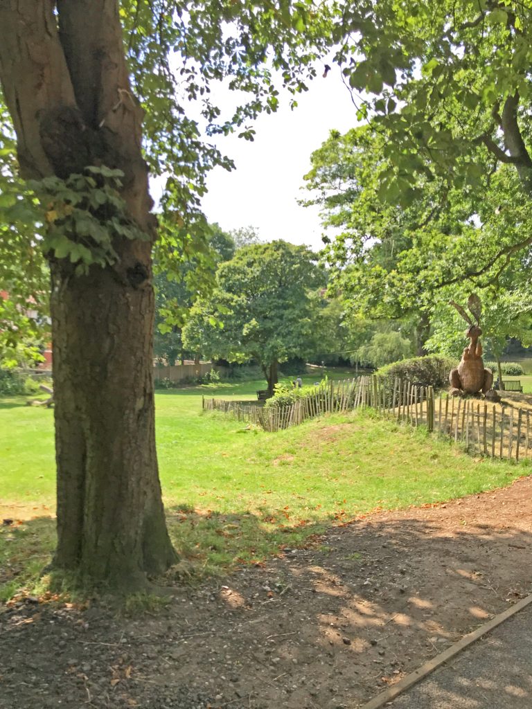 A photo of a mound covered in grass with a willow hare sculpture and lots of green trees