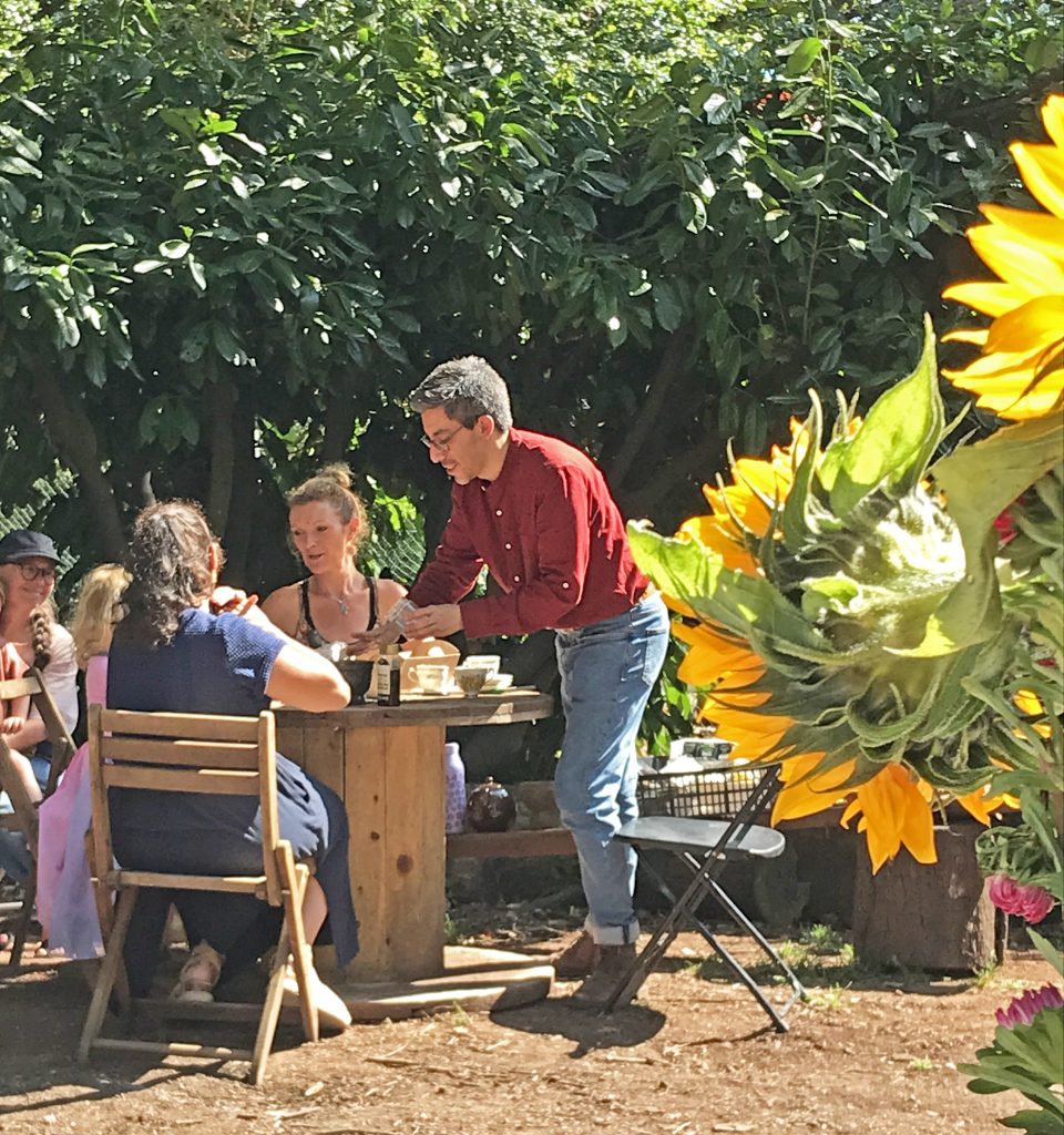 A photo of some people sat at a table in front of some bushes, with sunflowers in the foreground