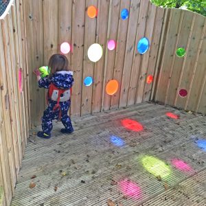 A photo of a toddler with her face up to the bright green porthole in a wooden structure, while other colours of porthole cast rainbow light nearby