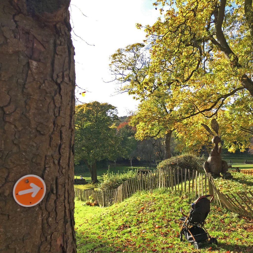 A photo of a grassy mound surrounded by autumn trees, with a willow hare sculpture and fence on top